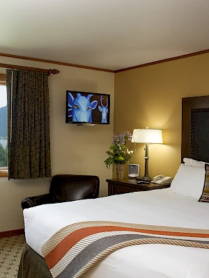 A cozy hotel room with a large bed, TV, and scenic window view of mountains and forest; warm lighting and decor complete the setting.