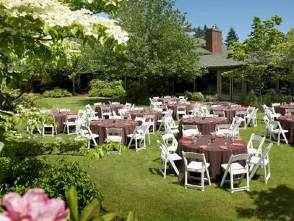 Outdoor garden setup with round tables covered in brown cloth and surrounded by white chairs, under a clear sky, surrounded by greenery.