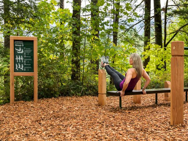 A person is exercising on a bench in an outdoor fitness area surrounded by trees, with an instruction board nearby.