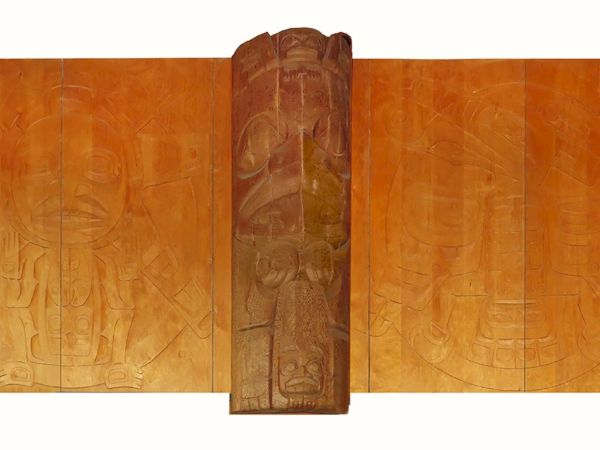 The image shows a wooden artwork with intricate carvings, featuring a central totem-like structure surrounded by engraved panels.