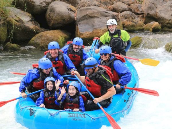 A group of people in helmets and life jackets rafting on a river, surrounded by rocks, smiling and waving in a blue inflatable raft.