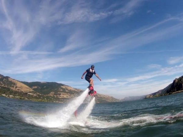 Two people are enjoying a flyboarding experience over a body of water, with mountains in the background under a clear sky.