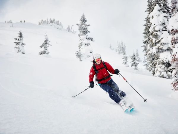 A person is skiing down a snowy slope surrounded by snow-covered trees, wearing a red jacket and helmet.