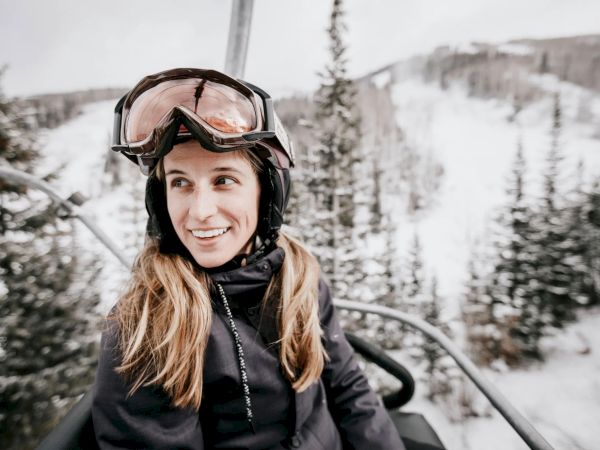A person wearing ski gear and goggles is sitting on a ski lift surrounded by snowy trees and slopes, smiling in the winter setting.