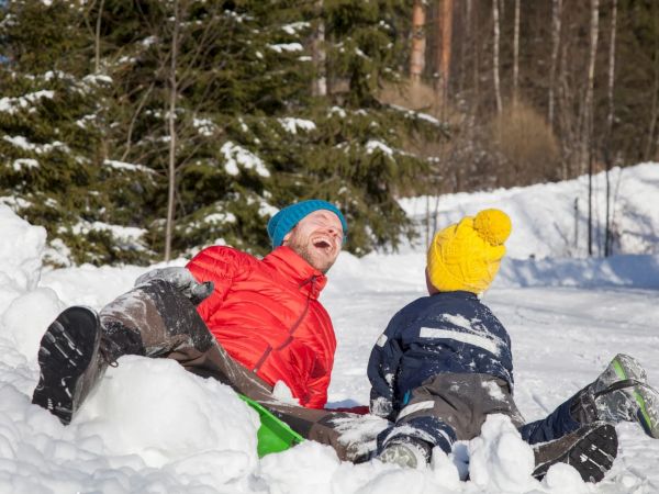 Two people in colorful clothing are playing and laughing in the snow against a backdrop of trees.