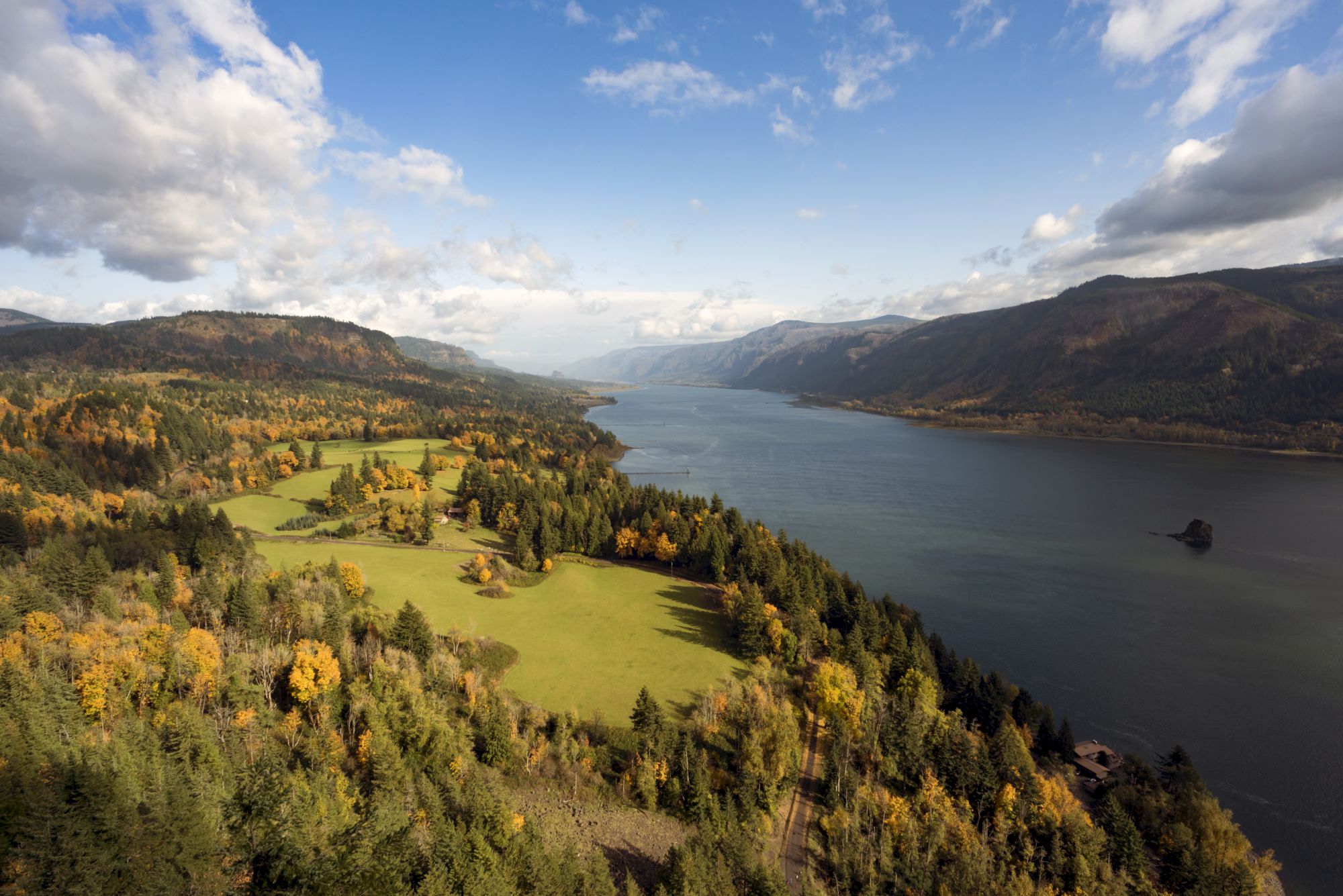 A scenic landscape featuring a river running through a valley, surrounded by lush forests and mountains under a partly cloudy sky.