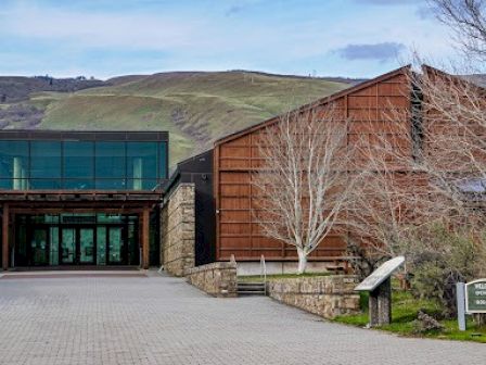A modern building with a glass entrance and rust-colored exterior is set against a hilly backdrop with bare trees in the foreground.