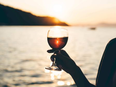 A hand holds a wine glass against a sunset backdrop over water, creating a serene and warm atmosphere.