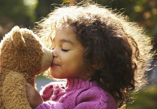 A child in a pink sweater kisses a teddy bear outdoors, with sunlight highlighting their curly hair in a peaceful setting.