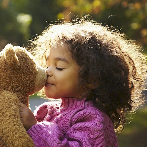 A child in a pink sweater kisses a teddy bear outdoors, with sunlight highlighting their curly hair in a peaceful setting.