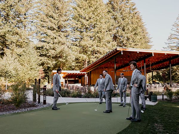 A group of people in suits stands on a putting green, with a wooden structure and trees in the background.