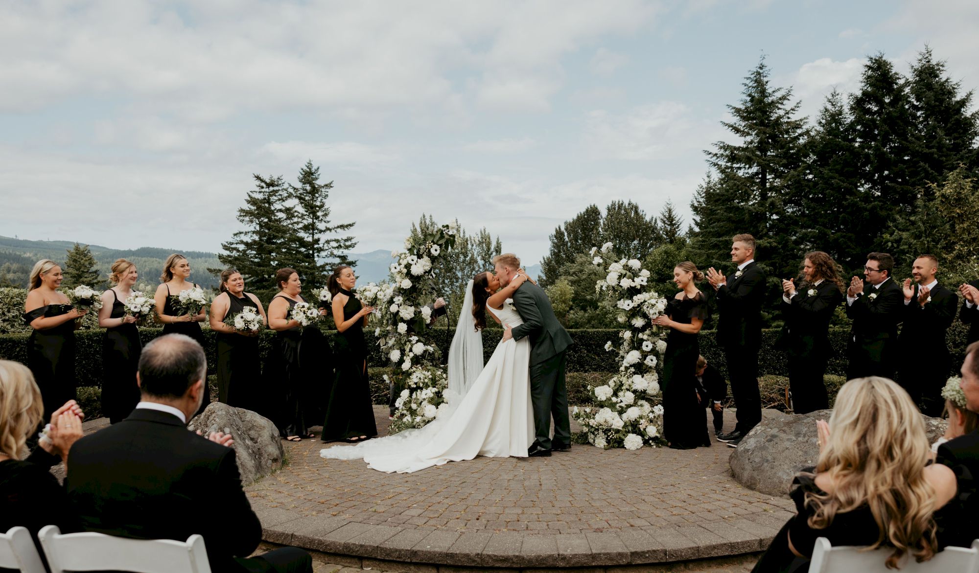 A couple is kissing at an outdoor wedding ceremony, surrounded by their wedding party and guests.