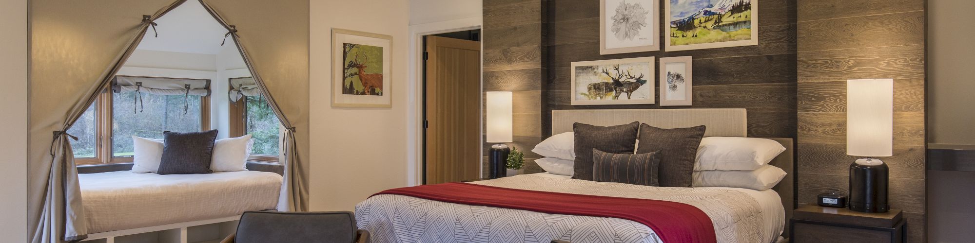 A cozy bedroom with a large bed, decorative pillows, two chairs, a rug, and a window nook in the corner, creating a relaxing atmosphere.