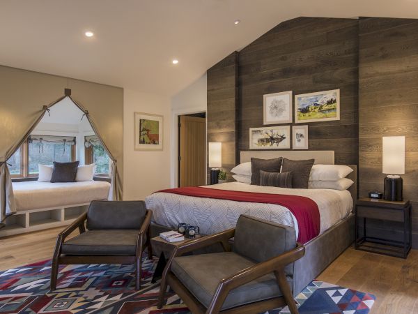 A cozy bedroom features a large bed, chairs, side tables, lamps, and artwork. A window seat with cushions is visible on the side.