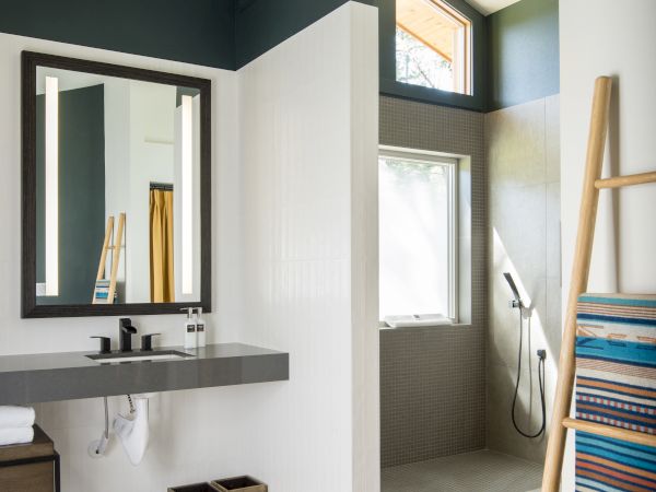 A modern bathroom with a mirror, countertop, walk-in shower, ladder with towels, and two storage baskets underneath.