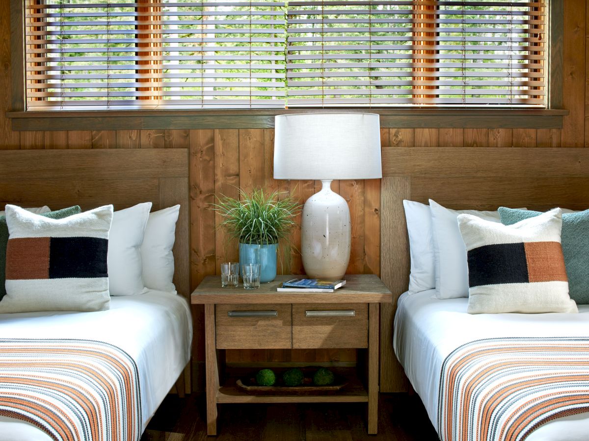A cozy bedroom with two beds, a central nightstand, a lamp, decorative pillows, and a plant, all against wood-paneled walls.