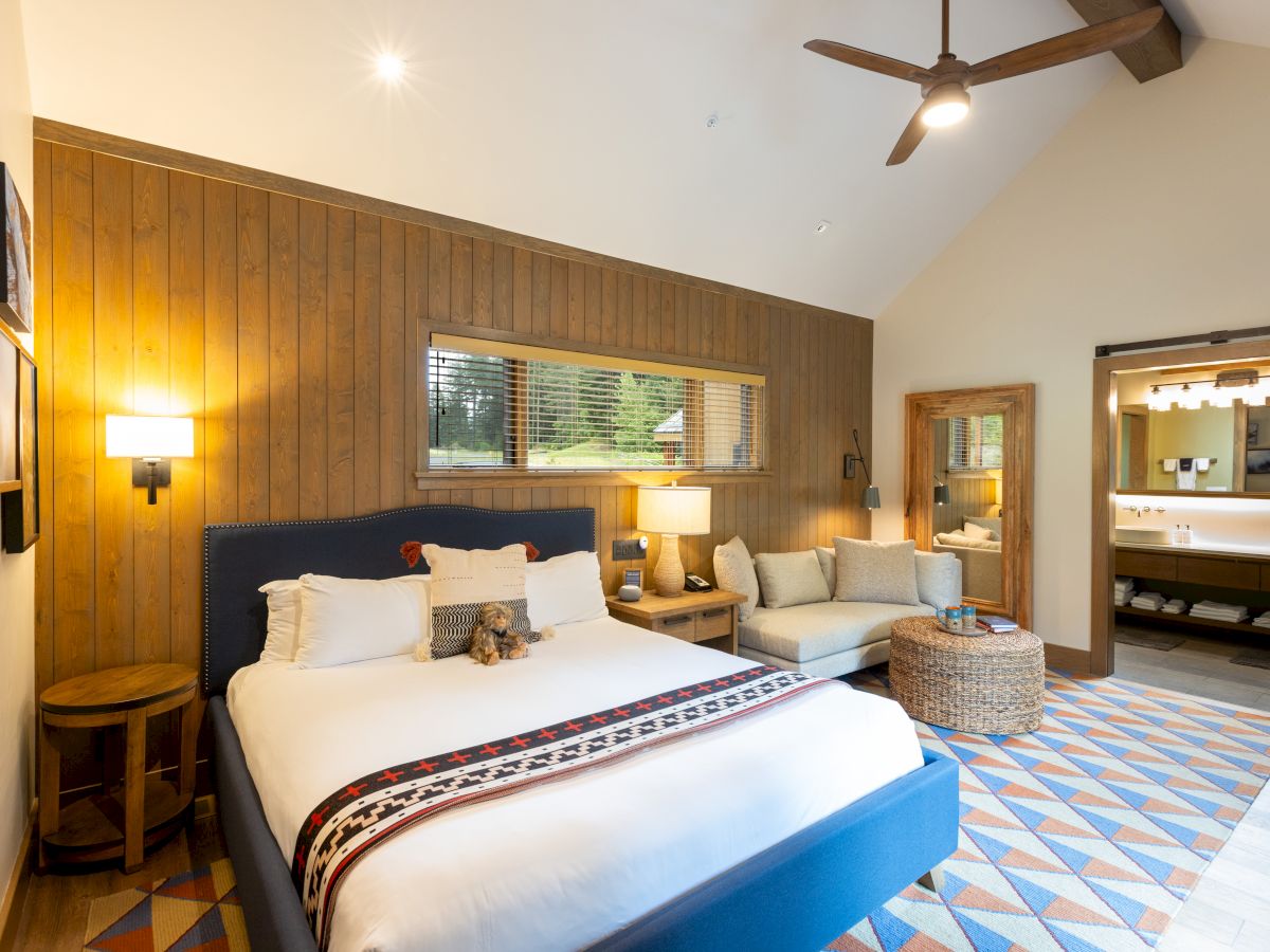 Cozy hotel bedroom with wood paneling, bed, sofa, ceiling fan, window, and an open bathroom view, with a small dog on the bed.
