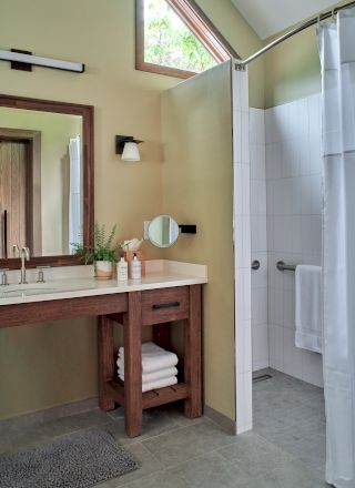 A bathroom with a wooden vanity, mirror, sink, and a shower area. Towels are folded on the shelf below. Natural light is present.