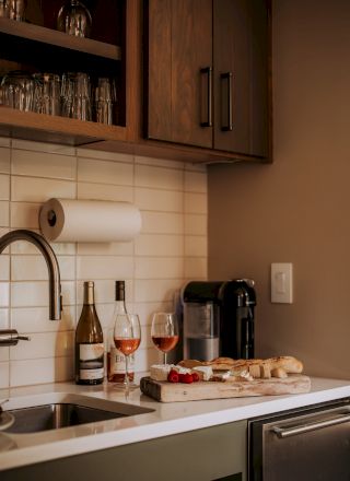 A kitchen counter with wine bottles, glasses, a coffee machine, and a cutting board with bread and cheese next to a sink.