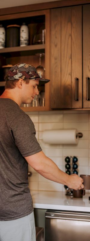 A person in a kitchen is making coffee, wearing a hat and T-shirt, standing beside a coffee machine.