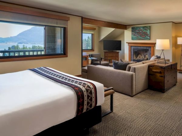 A cozy hotel room with a bed, seating area, fireplace, TV, and a window view of mountains and a lake.