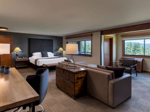 A spacious hotel room with a bed, couch, desk, and large windows offering a view of the greenery outside.