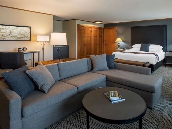 A modern hotel room with a large bed, gray sectional sofa, and a desk with a chair. There are lamps and a coffee table with books.