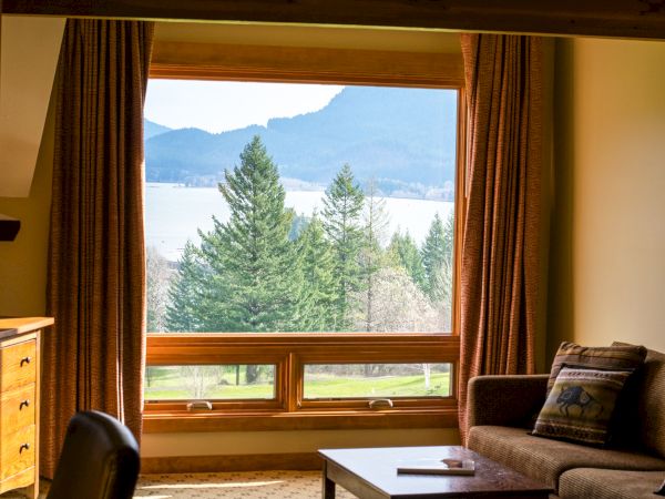 A cozy room with a large window view of trees, mountains, and a lake, featuring a sofa, cushions, and a wooden desk.