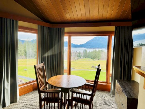 A cozy room with a wooden table and chairs, large windows showcase a scenic mountain and lake view, framed by green trees.