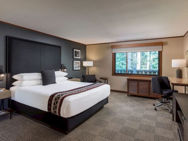 A hotel room with a bed, desk, chair, and window view of trees. There's modern decor, lamps, and framed pictures on the wall.