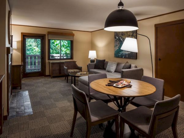A cozy living room with a round table, chairs, a gray sofa, floor lamp, and window offering a view of greenery.