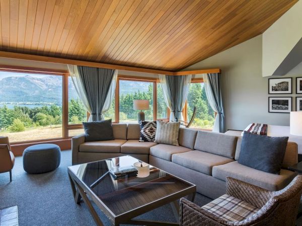 A cozy living room with large windows showing a scenic view, featuring a sectional sofa, glass coffee table, and framed photos on the wall.