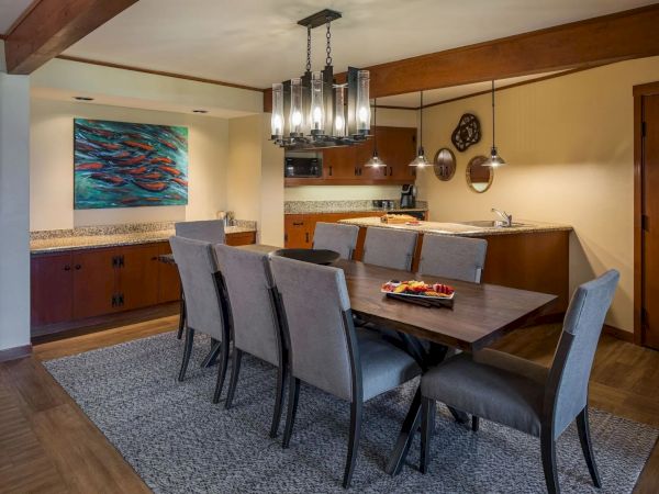 A dining room with a wooden table, gray chairs, a kitchen area, pendant lights, and a colorful fish artwork on the wall ends the scene.