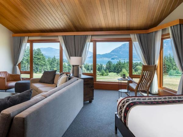 A cozy room with modern furniture, large windows showcasing a scenic view of trees, a lake, and mountains, under a wooden ceiling.
