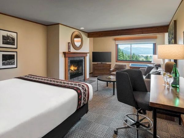 A cozy hotel room features a bed, fireplace, desk, and window view of trees and water, with modern furnishings and decor.