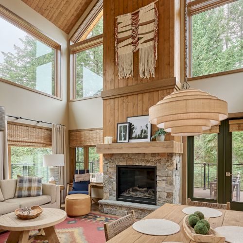 A cozy living room features large windows, a stone fireplace, wood accents, a woven wall hanging, and a stylish dining table.