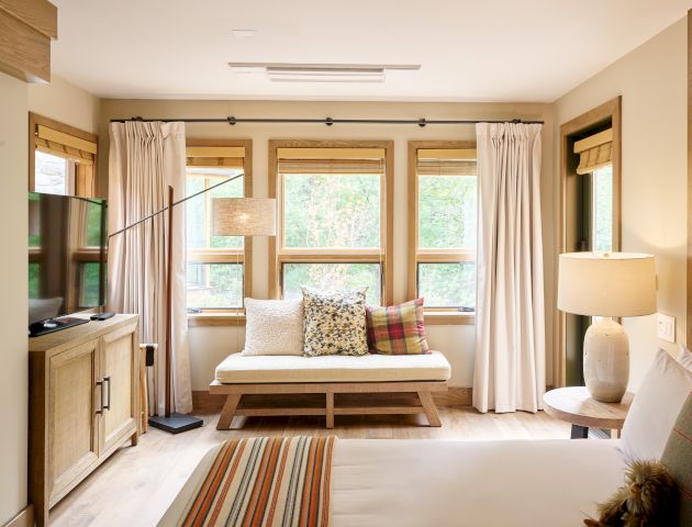 A cozy room with a bed, TV, cushioned bench, lamp, and windows with curtains, creating a warm, inviting atmosphere.