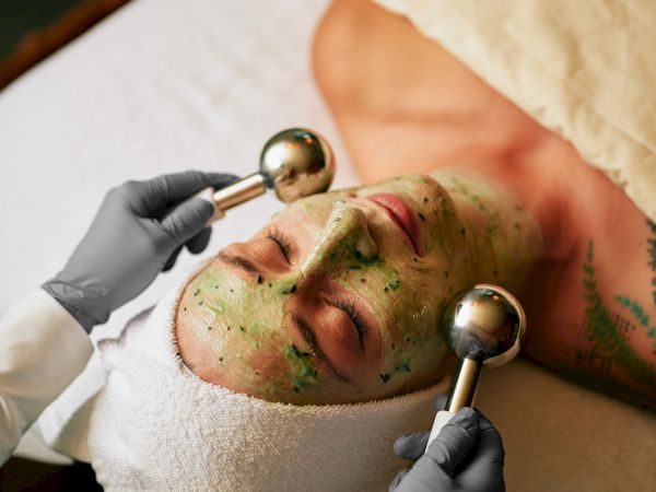 A person receiving a facial treatment with a green mask and massage wands, wearing a towel head wrap, on a spa table.