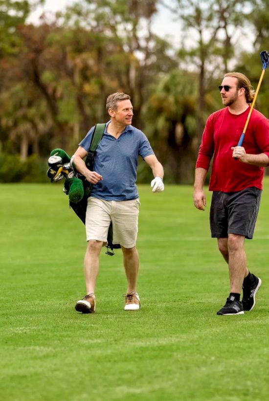 Two men walk on a sunny golf course, one carrying a bag and the other holding a putter, chatting as they stroll.