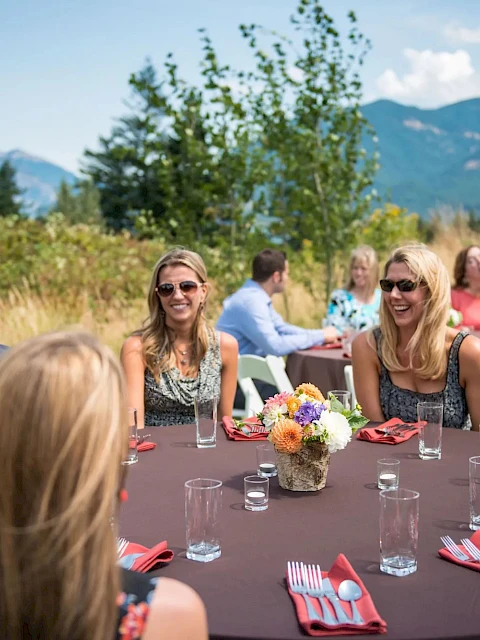 A group of people sitting at a round table outdoors, with a brown tablecloth, red napkins, and a mountain view in the background.