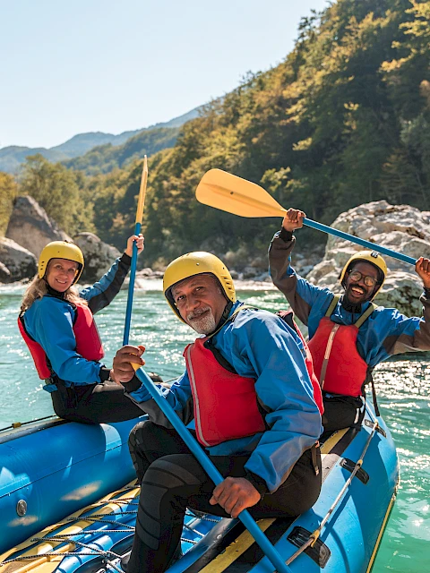 Three people in a blue raft with yellow helmets paddling on a bright river, forested canyons around, sunny day.