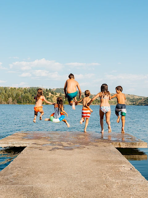 A family and friends jump off a wooden dock into a calm lake, sunny day, forested hills in the background.