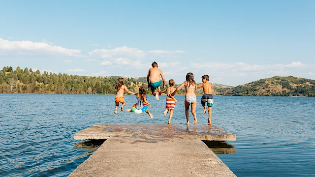 A family and friends jump off a wooden dock into a calm lake, sunny day, forested hills in the background.