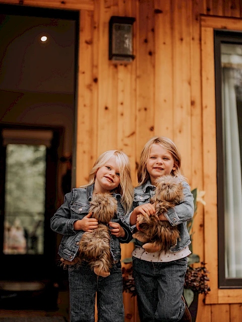 Two young girls stand outside a wooden house, each holding a fluffy puppy, smiling at the camera, with warm denim outfits and cozy vibes.