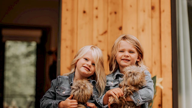 Two young girls stand outside a wooden house, each holding a fluffy puppy, smiling at the camera, with warm denim outfits and cozy vibes.