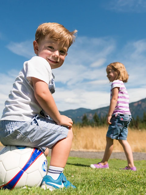 Two kids playing soccer outdoors: a smiling boy sits on a ball in the foreground while a girl walks in a sunny field with mountains in the background.