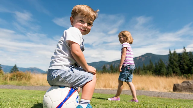Two kids playing soccer outdoors: a smiling boy sits on a ball in the foreground while a girl walks in a sunny field with mountains in the background.
