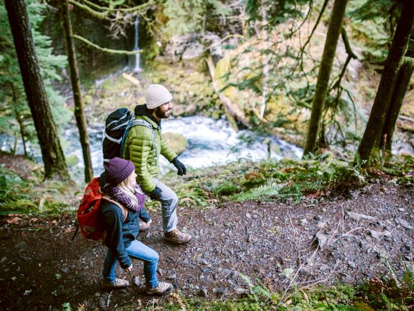Two people hiking on a forest trail with backpacks, enjoying the outdoors near a stream.