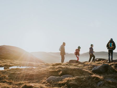 A group of four people hiking on a rocky landscape with backpacks, silhouetted against a bright sky and distant hills.