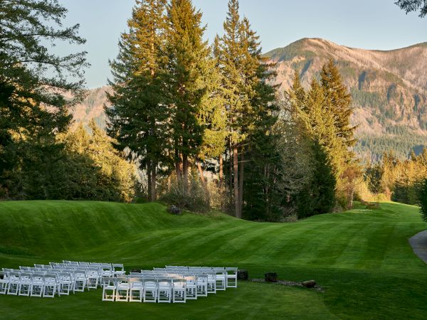 A grassy outdoor setting with rows of white chairs, surrounded by trees and mountains, likely prepared for an event or gathering.